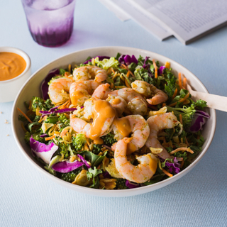 White bowl with roasted shrimp over super greens salad made with colorful cabbage, grains and herbs on a blue background with a purple glass in the background. 