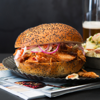 Side view of a barbecue chicken sandwich on a poppyseed bun with spicy potato salad in a dish next to it with a glass and a bottle of beer. 