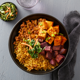 Black bowl with southern tofu, roasted beets and grains on a blue wooden table with a dark grey napkin and a small bowl of slaw on the side. 