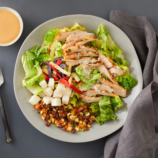 Southwestern chicken salad with chopped romaine lettuce, sliced chicken, tortilla strips, cotija cheese, and black beans with an orange dressing in a dish on the side and a grey linen napkin. 