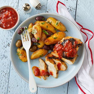 Sliced grilled Spanish chicken with roasted potatoes on a teal plate with a silver fork resting on top on a white wooden table with a red and white napkin on the side. 