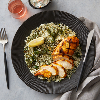 Spanakopita rice with spiced grilled chicken sliced into thin slices on a black round plate with a slice of fresh lemon on top with a fork and a knife on the side. 