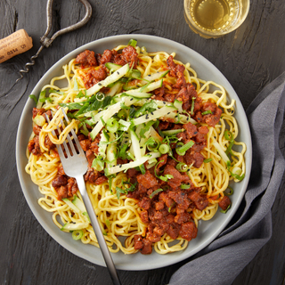 Spicy pork noodle bowl with long pasta, red sauce and topped with sliced green cucumbers with a fork on top in a round plate on a dark background. 