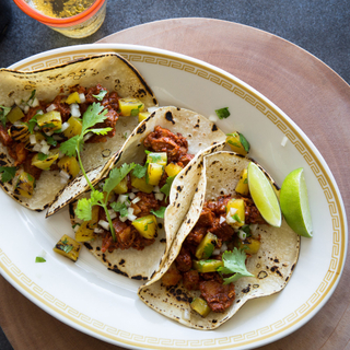 three spicy pork tacos on an oblong white plate with fresh lime, pineapple and cilantro