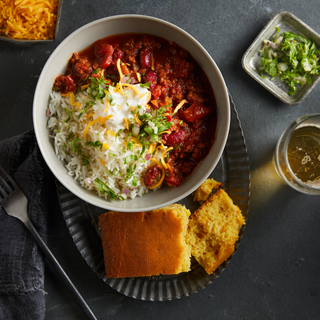 Spicy turkey chili topped with shaved cheese in a white bowl on a dark plate with a side of cornbread on a dark background with a glass of wine on the side and a fork in the corner. 