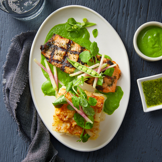 Spring salmon fillets with a square slice of potato gratin and bright green sauce on a white plate with raw vegetables for garnish on a dark wood background. 