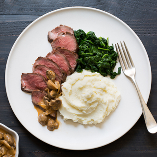 Steak Diane with sliced rare steak, mashed potatoes and wilted spinach on a white dinner plate on a dark wooden table with a silver fork on the plate. 