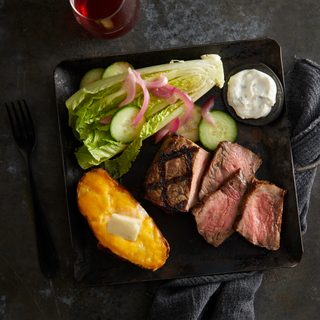 Steak house dinner with a half romaine heart, sliced grilled steak and a baked potato with a pat of butter on a black square plate on a dark background