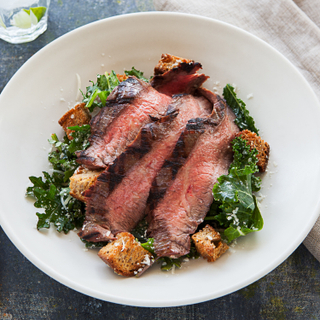 Salad with large slices of rare steak, kale and croutons on a white bowl with a white linen napkin in the background. 