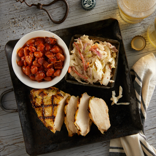 Summer BBQ chicken plate with grilled chicken sliced into three pieces on a black square plate with a bowl of bacon ranchero beans and a plate of coleslaw on a wooden table with a glass of beer. 