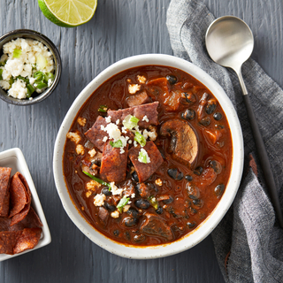 Portobello chili soup with corn tortilla chips on top and black beans in a white bowl on a blue wooden table with a grey towel and a spoon on the side. 