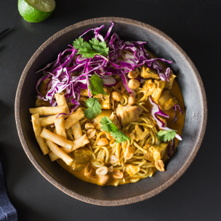 Thai chicken curry noodles with red cabbage, fresh cilantro in a black round bowl on a black background with a blue napkin and black chopsticks. 