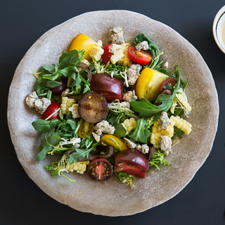 Tomato sweet corn salad with arugula, sliced tomatoes and cheese on a stone plate on a dark blue background with dressing on the side. 