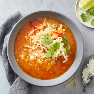 Red tortilla soup with sliced red peppers, fresh cilantro and cheese on top in a grey bowl with a grey napkin on a grey background. 