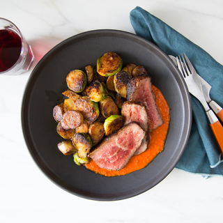sliced spanish-style trip tip with red romesco sauce, roasted potatoes and brussels sprouts in a black bowl