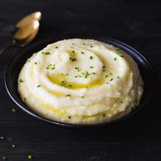 Black bowl of truffled mashed potatoes with sliced chives on top with two silver spoons in the background. 