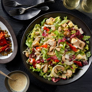 Tuscan tuna salad with white beans and dressing in a black round bowl on a black background with forks and plates and glasses of wine on the side. 