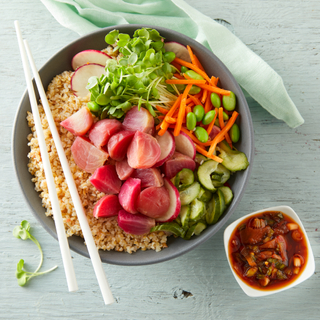 Vegan beet poke bowl with quinoa, roasted beets, sliced cucumbers, greens, radishes, edamame, carrots in a black bowl with dressing on the side and white chopsticks.