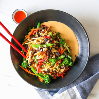 Vegan dan dan noodles with chili oil and fresh vegetables ina  black bowl with red chopsticks on a white background with a blue and white napkin. 