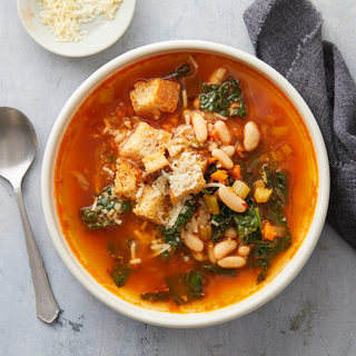 White bowl of white bean kale soup with cheese and croutons on a grey background with shredded cheese on the side. 