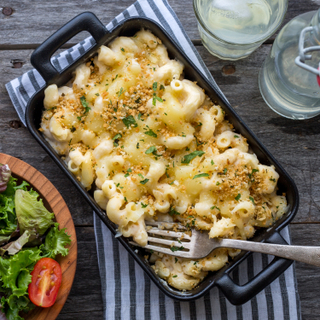 Dish of white cheddar mac and cheese in a cast iron pan next to a side salad in a wooden bowl with a grater on the side on a wooden table. 