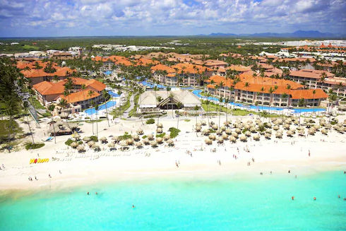Aerial view of a large tropical beach resort with orange roofs, winding pools, and a palapa-dotted white sand beach under a cloudy sky.