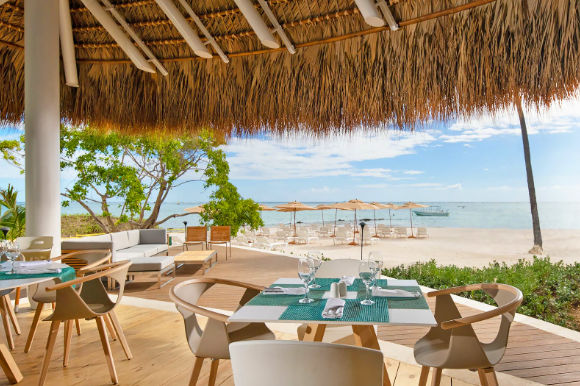 Open-air beachfront restaurant dining area under a thatched palapa roof overlooking a tropical white sand beach and calm ocean