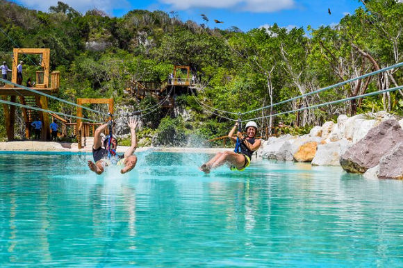 Two people splashing into a turquoise pool from a water zipline at a tropical adventure park