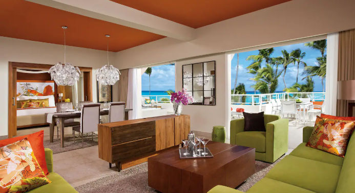 Luxury open-plan resort suite with living area, dining table, and a balcony overlooking palm trees and the ocean
