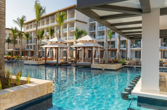Luxury resort swimming pool with private cabanas, a swim-up bar, palm trees, and modern hotel buildings.