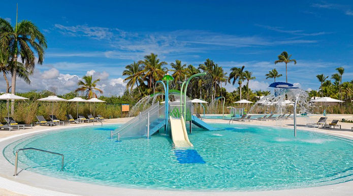 A children's resort splash pool featuring multiple water slides and spray features, surrounded by palm trees and lounge chairs under a blue sky.