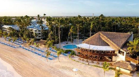 Aerial view of a beachfront resort in Punta Cana featuring a thatched-roof restaurant, swimming pool, palm trees, and blue lounge chairs on white sand
