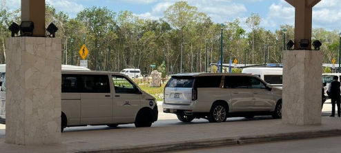 Lujosa SUV de transporte privado plateada y furgoneta de pasajeros blanca estacionadas en una zona de recogida de hotel bajo una marquesina, con vegetación y personal de servicio uniformado al fondo.