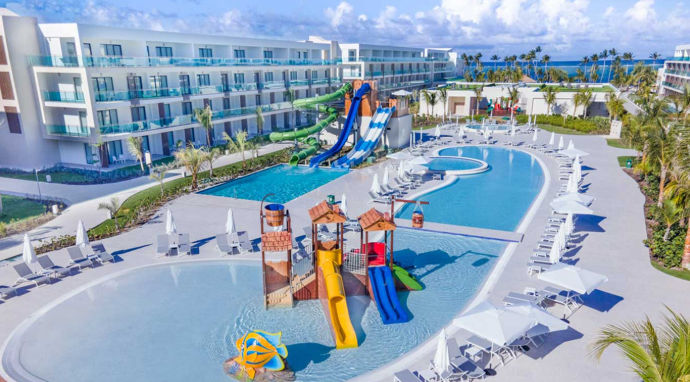 Aerial view of a tropical beach resort featuring a large swimming pool, a colorful kids water park, tall water slides, and white hotel buildings near the ocean