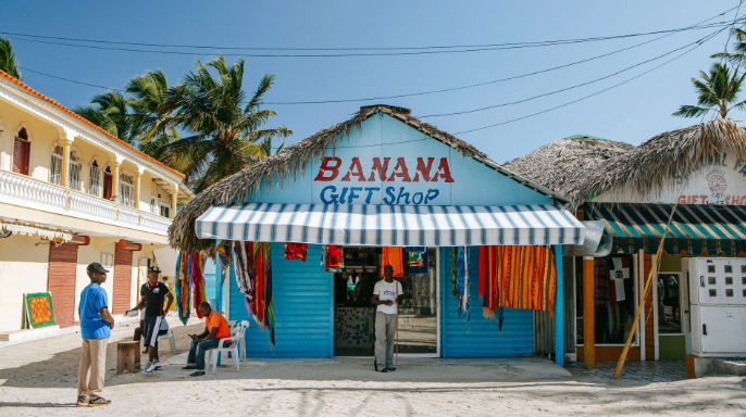 Fachada azul de la tienda Banana Gift Shop con techo de palma y artículos coloridos en una calle de arena