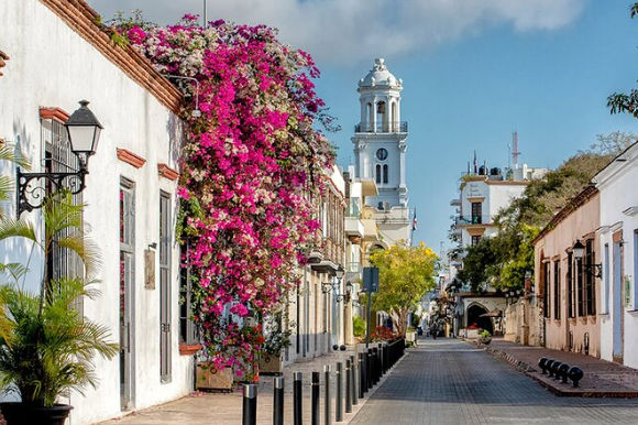 Hermosa calle colonial con fachadas blancas, coloridas enredaderas de flores rosas y una torre de reloj al fondo