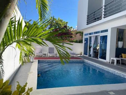 Exterior view of the Gaia Residence building, featuring a modern white facade with bright red accents, blue-tinted windows, and small palm trees along the front perimeter under a clear blue sky.