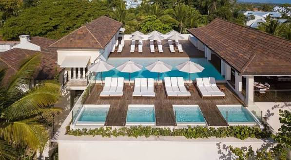 A wide-angle photograph of a luxurious building entrance featuring a decorative black metal portico supported by columns, an ivy-covered white facade with large windows and awnings, and a black luxury SUV parked on a patterned stone paver driveway under a clear blue sky with tropical trees.