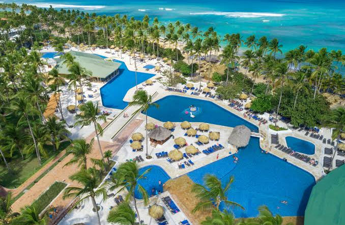 Aerial view of a hotel swimming pool with a white pedestrian bridge, surrounded by palm trees, lounge chairs, and with the ocean in the background.