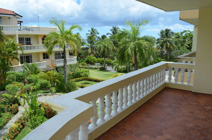 A detailed photo of a multi-story cream-colored apartment or resort building with numerous white-balustraded balconies and terracotta-tiled roofs, set within a lush tropical garden with many palm trees. A large, free-form swimming pool with bright blue water occupies the foreground, surrounded by a paved deck with lounge chairs and some people relaxing. To the right, there is a thatched-roof palapa covering a bar. The sky is clear and blue.