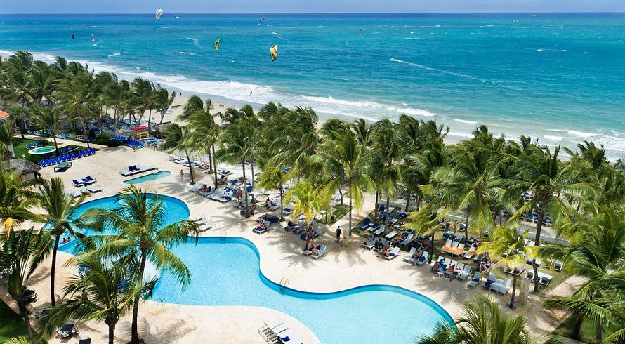 A view across a large blue resort swimming pool towards multi-story white hotel buildings with red tiled roofs and balconies, surrounded by palm trees, sun loungers, and patio tables under a clear sky.