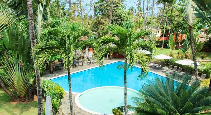 A tropical resort scene featuring a bright blue swimming pool with stone paving. Behind the pool is a multi-story terracotta-colored hotel building with a thatched roof section and wooden balconies, surrounded by lush palm trees and other greenery. White lounge chairs with umbrellas are lined up on the left, and a patio dining set is on the right under a blue, partly cloudy sky.