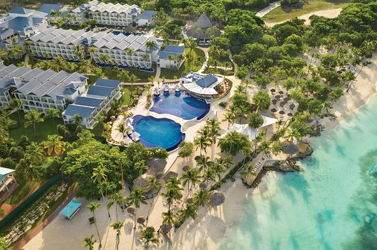An aerial photograph showing a luxurious tropical resort with several large, winding blue swimming pools surrounded by lounge chairs, white cabanas, and palm trees. A sandy beach with thatched palapas meets the blue ocean in the background under a cloudy sky.