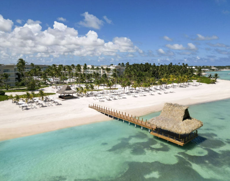 Vista aérea de un lujoso resort tropical en el Caribe con playa de arena blanca, aguas turquesas cristalinas y un muelle que lleva a una palapa sobre el agua