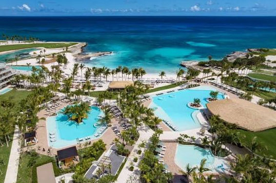 Vista aérea detallada de un complejo turístico costero de lujo, mostrando múltiples piscinas de color azul brillante rodeadas de tumbonas y densas palmeras, un edificio de hotel moderno a la izquierda, una playa de arena blanca y el océano turquesa en el fondo con un cielo azul.