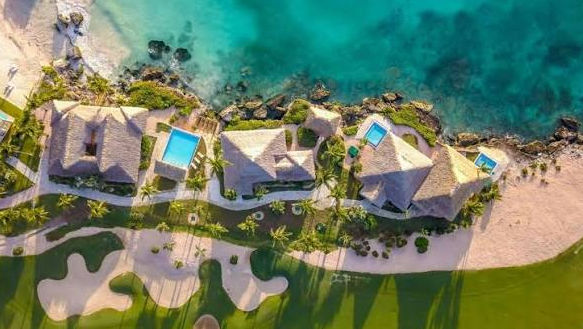 An aerial photograph of a line of luxurious thatch-roofed coastal villas nestled between a rocky turquoise ocean and a green golf course, with multiple private swimming pools, sandy beaches, and palm trees.