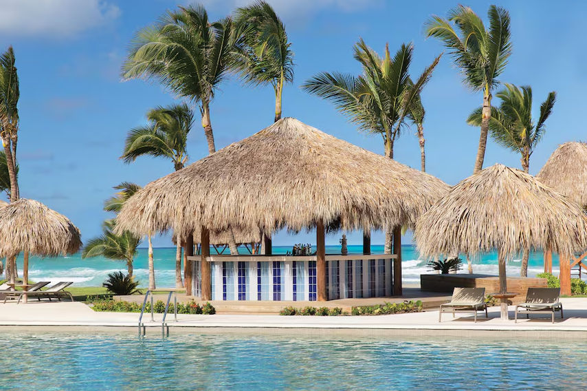 A scenic view of a tropical resort pool with a central thatched palapa bar and lounge chairs, overlooking the turquoise ocean and sandy beach under a blue sky with palm trees.