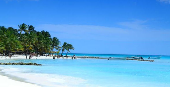 Scenic tropical beach with palm trees, white sand, crystal-clear turquoise water, small boats, and people wading under a clear blue sky.