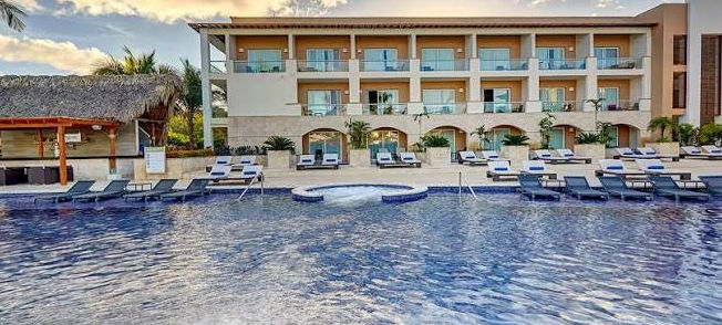 A modern resort pool area featuring lounge chairs, a thatched-roof bar, and a multi-story hotel building with balconies.