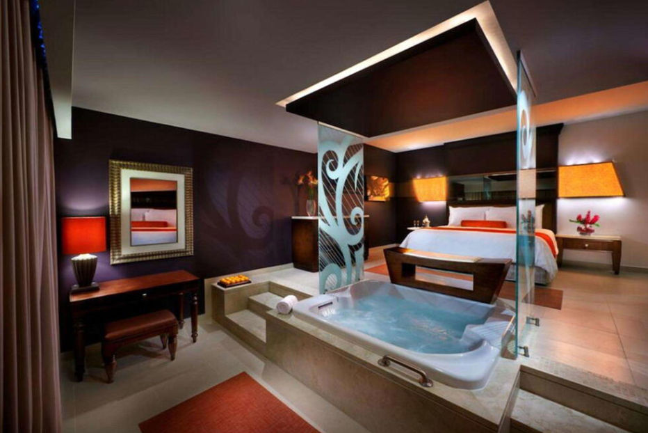 Modern luxury hotel suite featuring a private indoor jacuzzi in the foreground, decorative glass panels, and a bed with orange accents in the background.